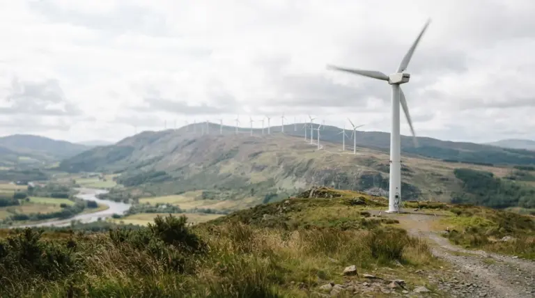 A wind turbine stands on a grassy hill overlooking a winding river and distant mountains with more turbines